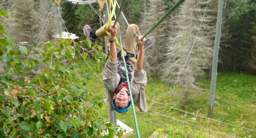 A person wearing safety gear and secured by ropes playfully hangs upside down on an obstacle on a high ropes course. 